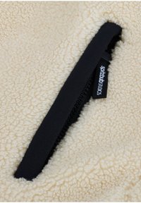 Fleece-Stoff in Off-White mit strukturierter Oberfläche, ausgestattet mit einer schwarzen Reißverschlusstasche mit einem Markenetikett, was die Funktionalität verbessert.