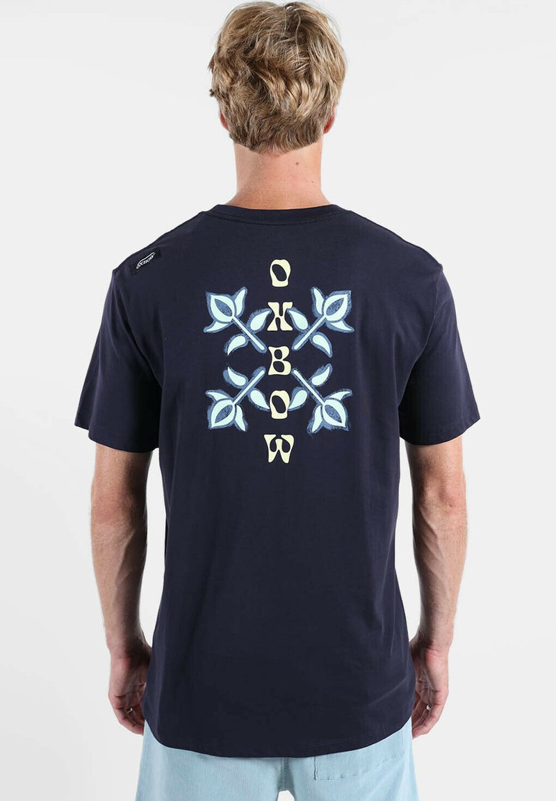 T-shirt en coton bleu marine avec un motif graphique de feuilles bleu clair et les lettres "OKBOW" au dos, manches courtes et col rond.