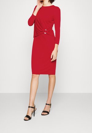 Femme portant une robe rouge cintrée à manches longues avec un détail de nœud torsadé à la taille, associée à des sandales noires à talons hauts.