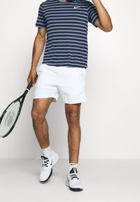 Muž v námořnickém pruhovaném tričku, bílých šortkách a bílých teniskách drží černou tenisovou raketu na jednobarevném bílém pozadí.