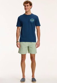 Marineblauw T-shirt met korte mouwen en palmboomgrafiek, gecombineerd met lichtgroene shorts. Model draagt grijze instapschoenen. Eenvoudig, casual ontwerp.