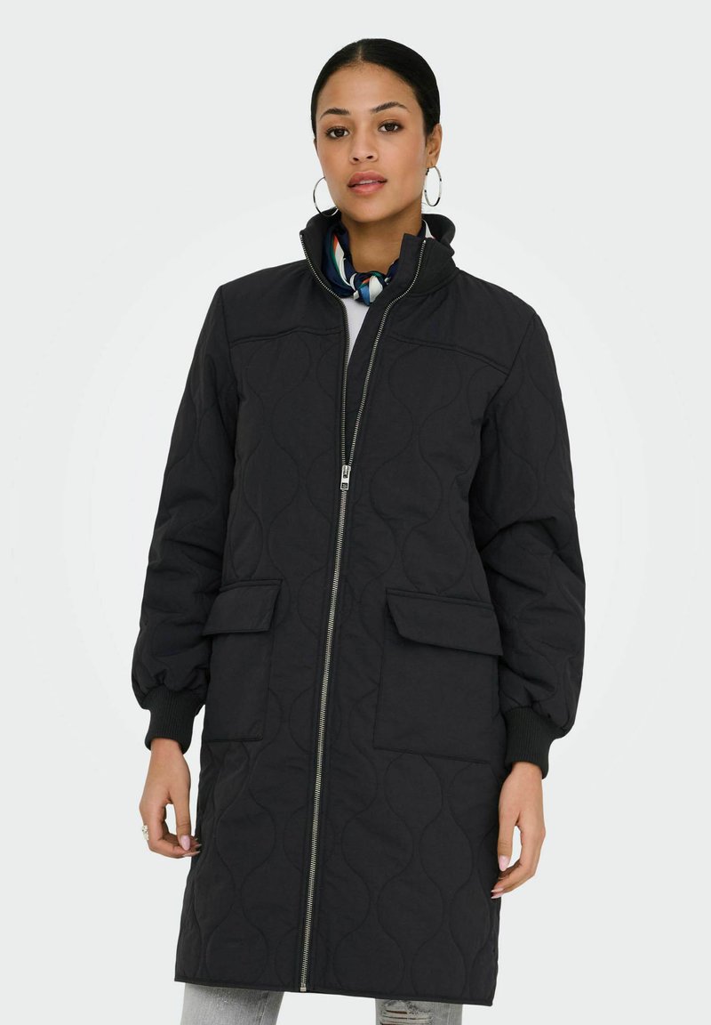 Femme portant un long manteau noir matelassé avec fermeture éclair frontale, poches à rabat, poignets côtelés et une écharpe à motifs autour du cou, sur un fond uni.