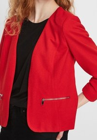 Red textured blazer features a fitted design with a scoop neck, zippered pockets, and three-quarter sleeves. Styled over a black shirt.