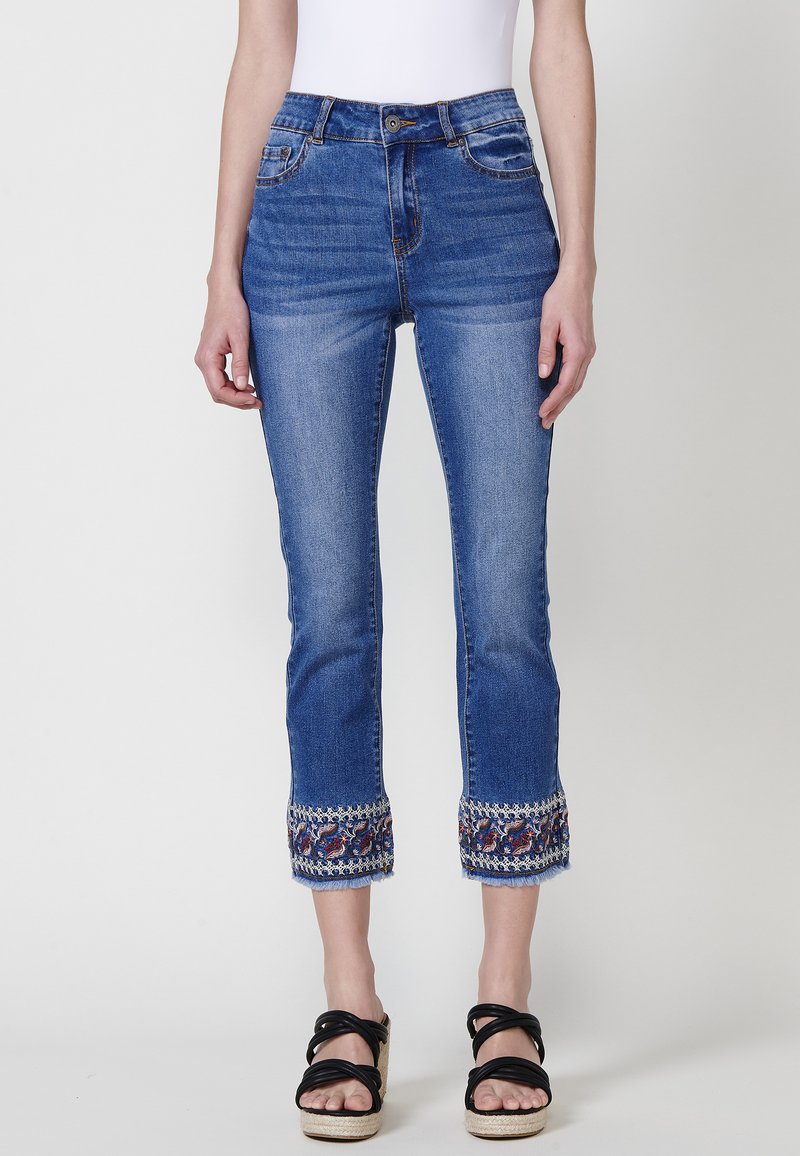 Blå denim jeans i cropped modell med broderade fållar. Hög midja, fem fickor, lätt blekning på benen och fransade kanter vid bensluten.