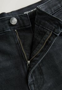 Jean en denim noir avec fermeture éclair et bouton ouverts, montrant l’étiquette « Regular Fit » sur la ceinture à l’intérieur.