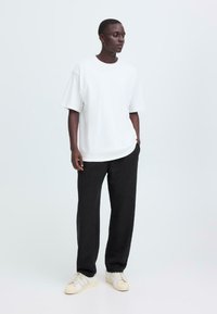 Weißes, übergroßes T-Shirt kombiniert mit schwarzen Sweatpants, mit einer lässigen Passform. Das Model trägt strukturierte Sneakers mit hellen Akzenten.