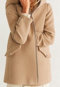 Manteau en laine beige avec capuche, fermeture éclair, poches à rabat et manches à boutons. Texture douce et silhouette cintrée.