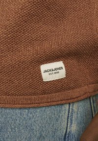 Barna kötött anyag texturált mintával; az oldal hemnél egy fehér címke található, rajta a "JACK&JONES EST.1990" felirattal. Farmer látható mögötte.