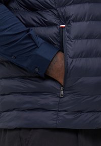 Gilet matelassé navy avec un design en quilté et une poche zippée. Texture lisse et brillante ; associé à une chemise boutonnée bleu foncé en dessous.