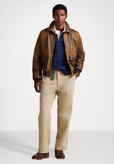 Bruine leren bomberjack, marineblauwe polo, beige relaxte broek met slijtage, bruine schoenen en een gestreepte riem.