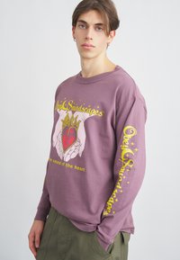 Ilgų rankovių levandų spalvos džemperis su grafiniu spausdinimu, kuriame pavaizduoti rankos laikantys širdį, geltonas tekstas ir žvaigždžių akcentai ant rankovių.