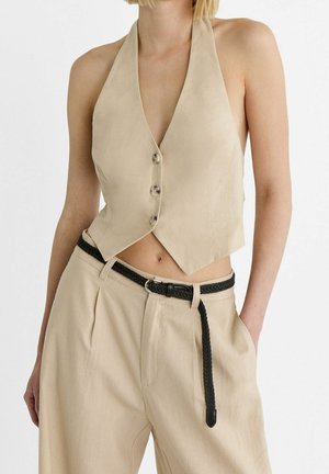 Vrouw draagt een beige mouwloze haltervest met knopen en bijpassende broek met een zwarte gevlochten riem, staand met één hand in de zak.