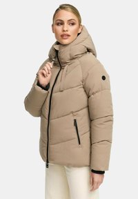 Vrouw die een beige pufferjack met een hoge kraag en capuchon draagt, naar de zijkant kijkt en de ritsstopper bij haar kin vasthoudt.