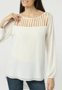 Mujer que lleva una blusa blanca de manga larga con rayas cortadas verticalmente en el pecho, combinada con jeans de color azul oscuro.