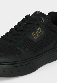 Μαύρο αθλητικό παπούτσι EA7 Emporio Armani με λεπτομέρειες από δίχτυ και δέρμα, μαύρα κορδόνια και χρυσό λογότυπο στο πλάι και στη γλώσσα.
