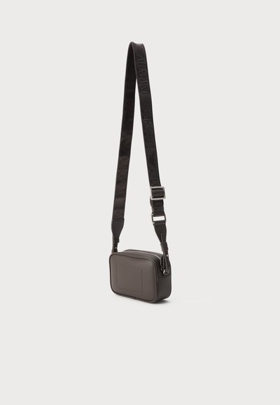 Μικρή σκούρα καφέ δερμάτινη τσάντα crossbody με φαρδιά ρυθμιζόμενη τιράντα με ανάγλυφο όνομα μάρκας και ασημένια μεταλλική αγκράφα.