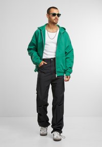 Groene zip-up hoodie met grote zakken, gedragen over een wit tanktop; gecombineerd met zwarte cargo broek met trekkoorddetails en sneakers.