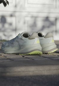 Light gray running shoes with a textured upper, green accents, and a cushioned sole. Features three stripes on the sides and a reinforced heel.
