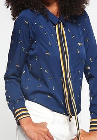 Mörkblå blus med krage, prydd med ett mönster av små gula och gröna fåglar, och randiga manschetter med guld- och vittoner.