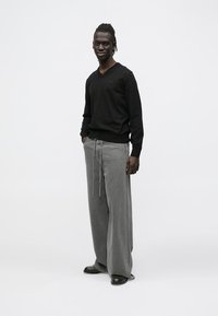 Férfi, aki zsebébe dugott kézzel áll, fekete, hosszú ujjú V-nyakú pulóvert, széles szárú szürke nadrágot és fekete cipőt visel, fehér háttér előtt.