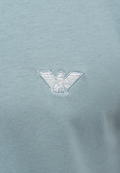 Svijetlo plava pamučna polo majica s suptilnim izvezenim logotipom orla u bijeloj boji na lijevoj strani grudi, s glatkom teksturom.