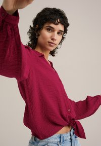 Magenta košulja s dugim rukavima i teksturiranim materijalom, zavezanog čvora u struku, u kombinaciji s svijetloplavim trapericama.