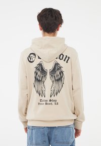 Felpa beige con cappuccio e coulisse, presenta una grafica nera a forma di ali e testo sul retro. Il materiale sembra morbido con una vestibilità comoda.