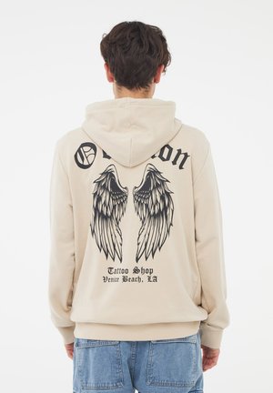 Felpa beige con cappuccio e coulisse, presenta una grafica nera a forma di ali e testo sul retro. Il materiale sembra morbido con una vestibilità comoda.