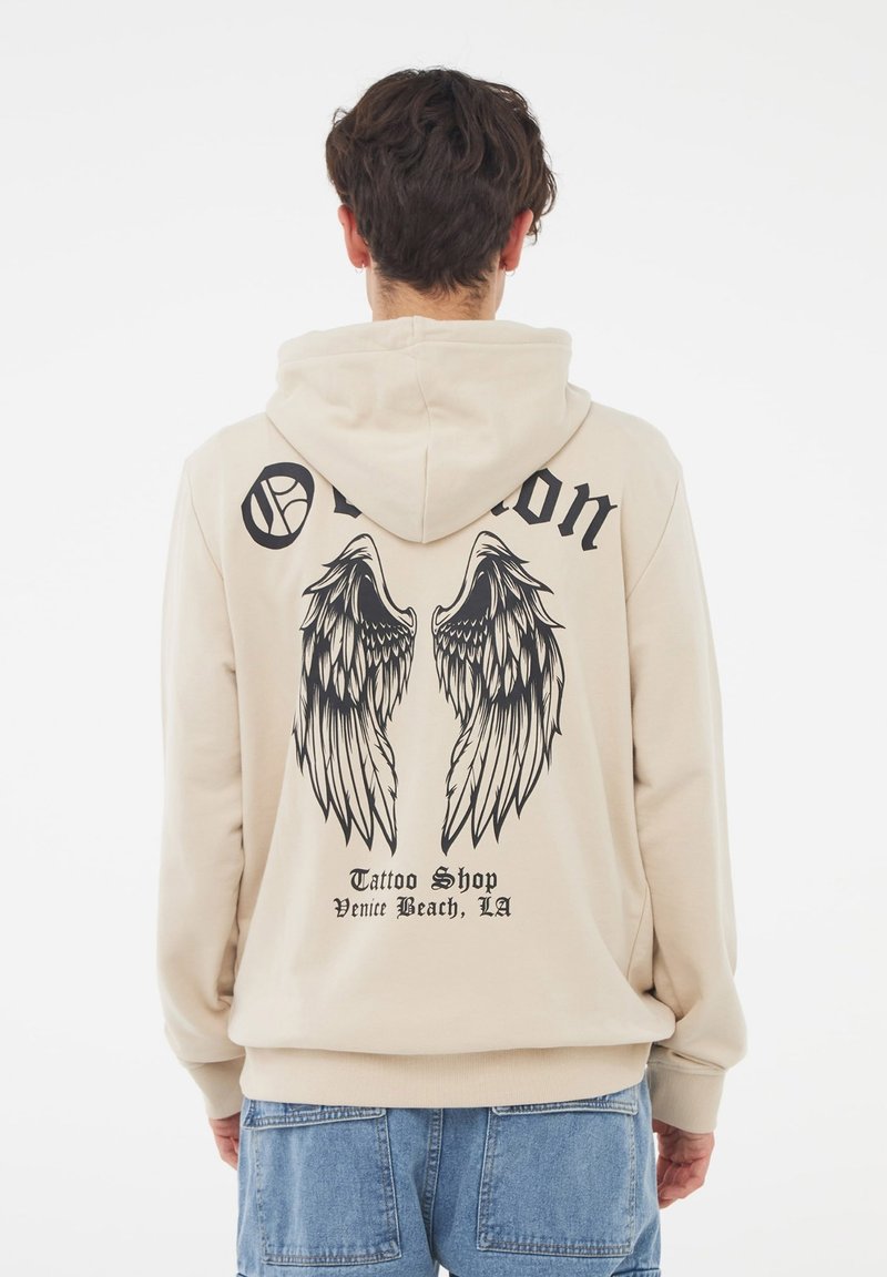 Felpa beige con cappuccio e coulisse, presenta una grafica nera a forma di ali e testo sul retro. Il materiale sembra morbido con una vestibilità comoda.