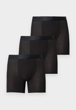 Høj, muskuløs mand stående barfodet mod en ensfarvet baggrund iført sorte boxershorts, vendt let til siden med et neutralt udtryk.