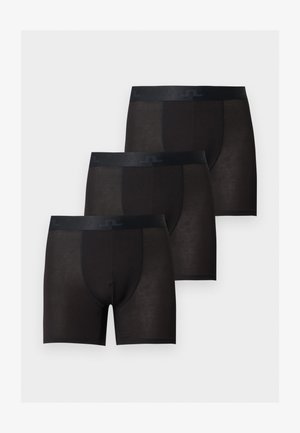 Homme grand et musclé, debout pieds nus sur un fond uni, portant un boxer noir, légèrement tourné de côté avec une expression neutre.