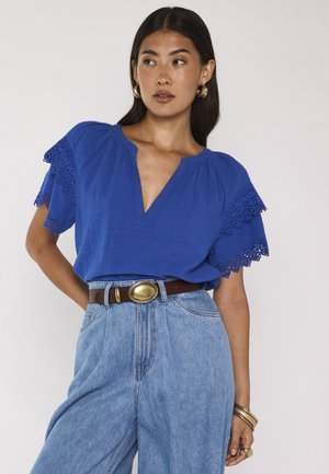 Donna che indossa una camicetta blu con maniche in pizzo e jeans azzurri a vita alta, accessoriata con orecchini a cerchio dorati, cintura, braccialetti e anelli.