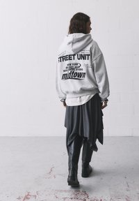 Haut à capuche gris avec un logo et un texte dans le dos, superposé à une jupe asymétrique gris foncé. Des bottes jusqu'aux genoux noires complètent la tenue.