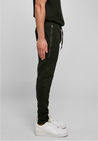 Pantalons de jogging noirs en tissu doux, avec une coupe ajustée, des poches zippées et une taille élastique, associés à des baskets blanches.