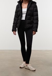 Czarna kurtka puchowa z kapturem, z zapięciem na zamek i elastycznymi sznurkami po bokach. Noszona z białą bluzką na ramiączkach, czarnymi legginsami i beżowymi sneakersami.