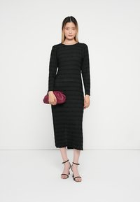 Vestido preto com mangas longas e riscas horizontais texturizadas. Complementado por uma mala de matiz burgundy e sandálias de tiras pretas.
