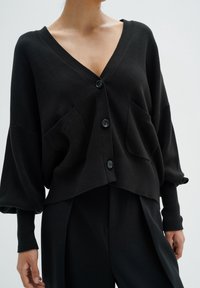 Černý zkrácený cardigan z měkké látky, s výstřihem do V, čtyřmi černými knoflíky a dvěma předními kapsami. Hladká textura a uvolněný střih.