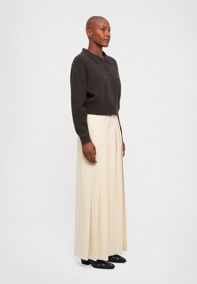 Personne debout portant un pull à boutons brun foncé, un pantalon large plissé beige et des chaussures noires à bout pointu sur un fond blanc uni.