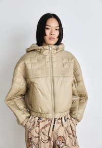 Beige quilted puffer jacket with a hood, zip front, and structured pattern detailing on shoulders. Styled with patterned trousers.