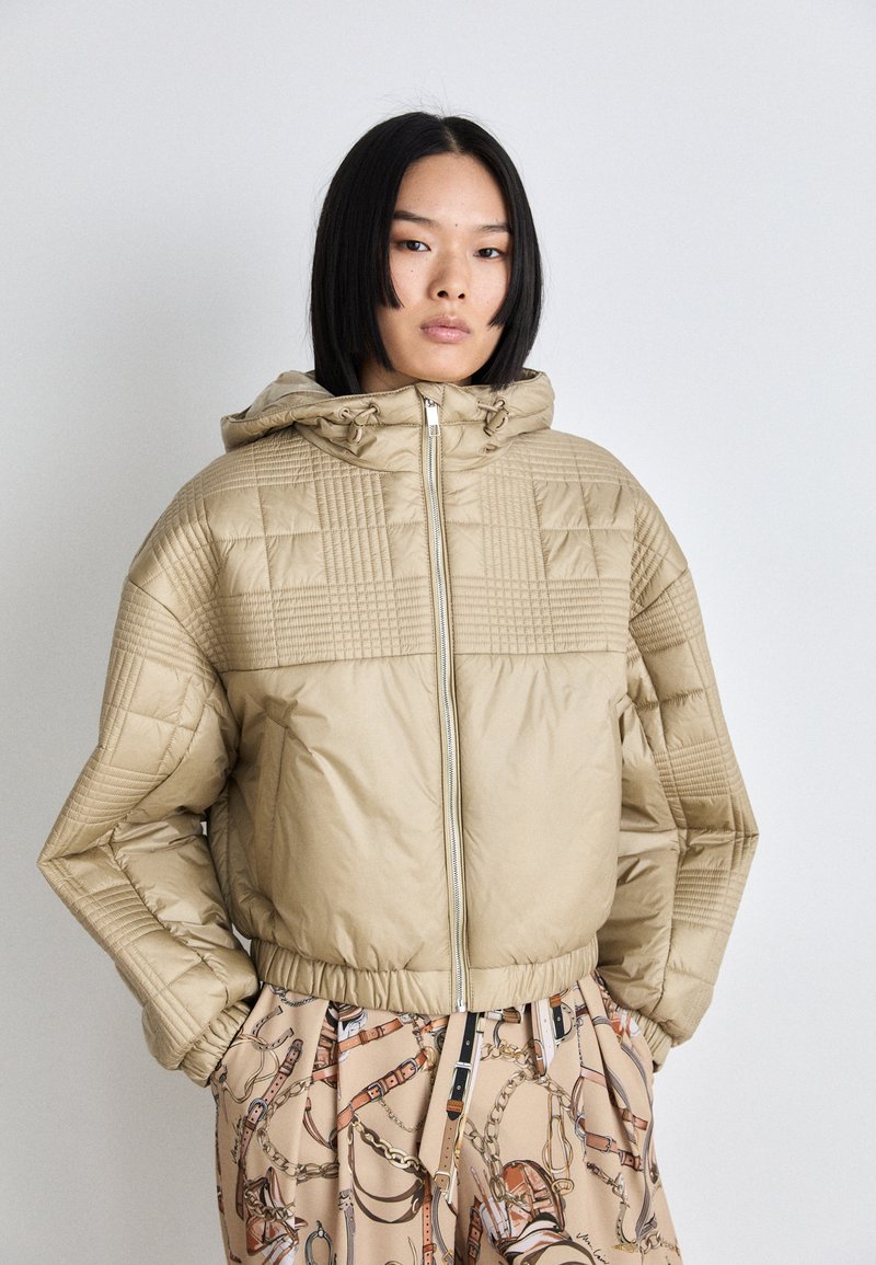 Beige quilted puffer jacket with a hood, zip front, and structured pattern detailing on shoulders. Styled with patterned trousers.