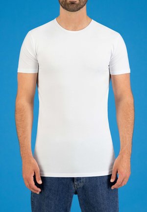 Witte T-shirt met korte mouwen en ronde hals, slim fit ontwerp en een gladde textuur, gecombineerd met blauwe denim jeans tegen een blauwe achtergrond.