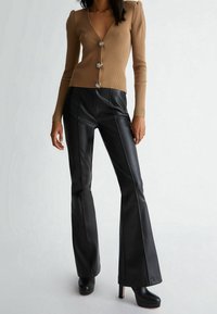 Gerippte braune Strickjacke mit tiefem V-Ausschnitt und drei silbernen Knöpfen, kombiniert mit schwarzen, ausgestellten Hosen aus Kunstleder mit Seitennähten.