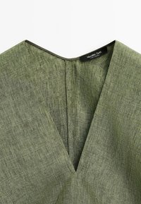 Abito in lino verde con scollatura a V, caratterizzato da una sottile trama e cuciture visibili; etichetta con il nome del marchio sulla parte posteriore della scollatura.
