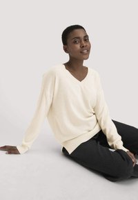 Cremefarbenes Strickpullover mit V-Ausschnitt, überschnittenen Schultern und gerippten Bündchen, kombiniert mit lockeren schwarzen Hosen, sitzend vor einem neutralen Hintergrund.