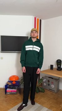 Grønn hettejakke med glidelås og to hvite horisontale striper, svarte bukser og mørke sko. Enkel design og avslappet passform.