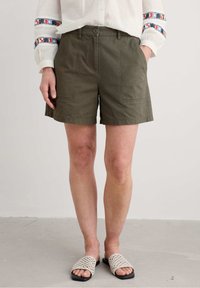 Olivgröna shorts tillverkade av bomullsblandning, med en rak framsida, två sidofickor och knappstängning. Pairade med vävda sandaler.