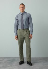 Helles graues Hemd mit Knöpfen, dunkler grauer Krawatte, olivgrüne Hose und schwarze Schuhe, alles in einer neutralen Umgebung mit sanften Texturen.