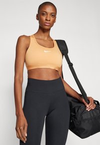 Nike oranžs sporta krūšturis ar baltu logo, savienots ar augsto vidukli melnajām legingām. Modelis tur melnu sporta somu ar plecu siksnu.