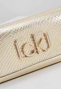 Kultainen, teksturoitu tekonahkainen clutch-laukku, jossa on keskiössä oleva korostettu metallilogo. Pinta on sileä ja siinä on hienovarainen matelijakuosis.