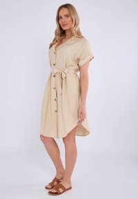 Abito camicia beige con maniche corte e vita accentuata da una cintura. Presenta un orlo leggermente svasato e un tessuto testurizzato. Indossato con sandali marroni.